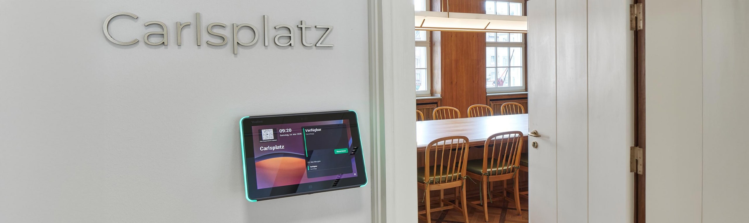 Blick in einen Besprechungsraum einer Anwaltskanzlei. Links neben der Tür ein Ipad das die Verfügbarkeit anzeigt. Aufschrift Carlsplatz an der Wand